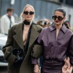 Warsaw Fashion Week 2025 - czy Warszawa będzie miała swój tydzień mody? Data, lokalizacja, szczegóły. Na zdjęciu Dwie stylowo ubrane kobiety idą ramię w ramię ulicą podczas Warsaw Fashion Week. Po lewej blondynka w oversize’owym, oliwkowym płaszczu o szerokich ramionach, z dużymi okularami przeciwsłonecznymi, złotymi kolczykami i czarną, skórzaną torebką trzymaną w dłoni. Pod płaszczem ma głęboko rozpiętą marynarkę i jedwabną chustkę zawiązaną na szyi. Po prawej brunetka ze spiętymi gładko włosami, w dużych okularach przeciwsłonecznych, ciemnofioletowej kurtce z zamkiem i dopasowanej spódnicy, trzyma pudroworóżową, marszczoną torebkę.