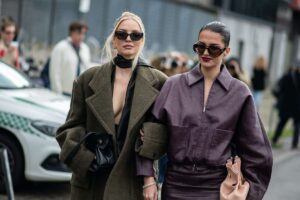 Warsaw Fashion Week 2025 - czy Warszawa będzie miała swój tydzień mody? Data, lokalizacja, szczegóły. Na zdjęciu Dwie stylowo ubrane kobiety idą ramię w ramię ulicą podczas Warsaw Fashion Week. Po lewej blondynka w oversize’owym, oliwkowym płaszczu o szerokich ramionach, z dużymi okularami przeciwsłonecznymi, złotymi kolczykami i czarną, skórzaną torebką trzymaną w dłoni. Pod płaszczem ma głęboko rozpiętą marynarkę i jedwabną chustkę zawiązaną na szyi. Po prawej brunetka ze spiętymi gładko włosami, w dużych okularach przeciwsłonecznych, ciemnofioletowej kurtce z zamkiem i dopasowanej spódnicy, trzyma pudroworóżową, marszczoną torebkę.
