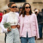 Zara letnia wyprzedaż 2025 - kiedy start? Na zdjęciu dwie eleganckie kobiety stoją razem na tle miejskiego krajobrazu, ubrani w modne tweedowe marynarki. Jedna z nich ma białą, krótką marynarkę, białe topy, dżinsy z wysokim stanem oraz perłowe akcesoria i trzyma zieloną torebkę. Druga ma na sobie jasnoróżową marynarkę z perłowymi guzikami, niebieskie jeansy i ciemne okulary przeciwsłoneczne. Obydwie noszą stylowe okulary przeciwsłoneczne i wyglądają pewnie, z modnym, paryskim akcentem.