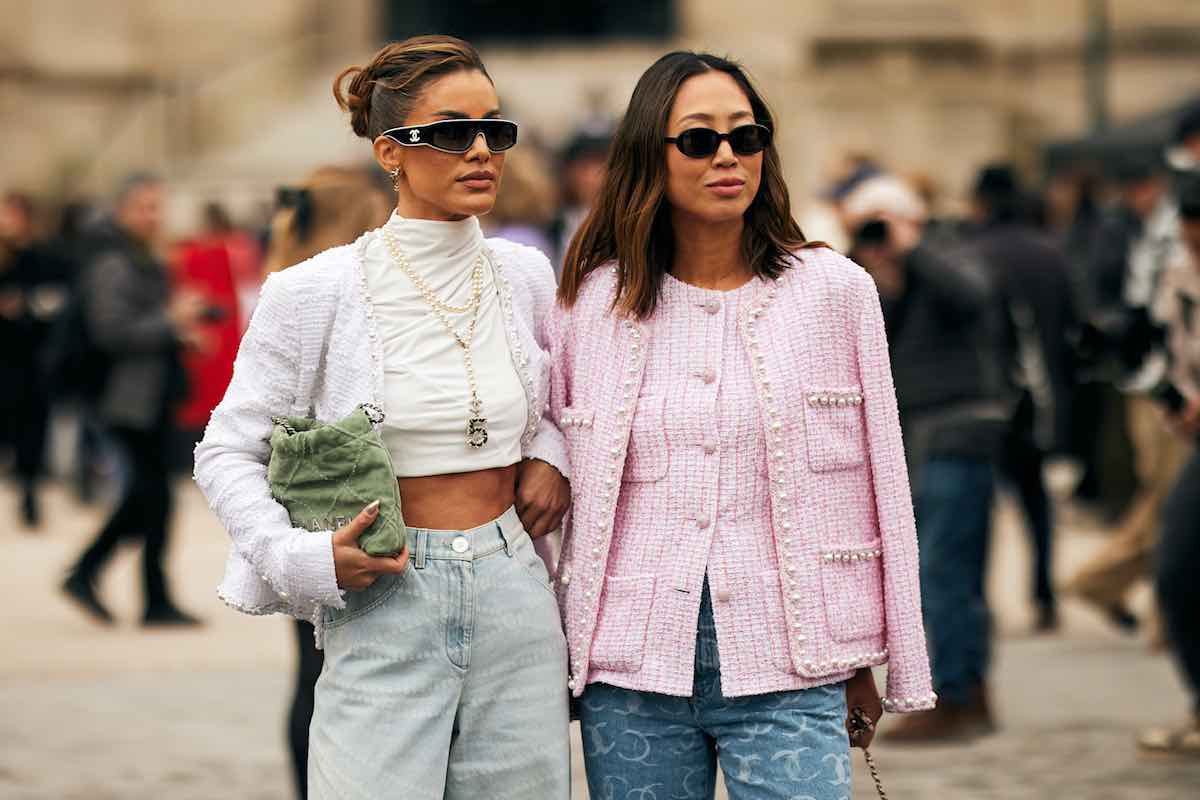 Zara letnia wyprzedaż 2025 - kiedy start? Na zdjęciu dwie eleganckie kobiety stoją razem na tle miejskiego krajobrazu, ubrani w modne tweedowe marynarki. Jedna z nich ma białą, krótką marynarkę, białe topy, dżinsy z wysokim stanem oraz perłowe akcesoria i trzyma zieloną torebkę. Druga ma na sobie jasnoróżową marynarkę z perłowymi guzikami, niebieskie jeansy i ciemne okulary przeciwsłoneczne. Obydwie noszą stylowe okulary przeciwsłoneczne i wyglądają pewnie, z modnym, paryskim akcentem.