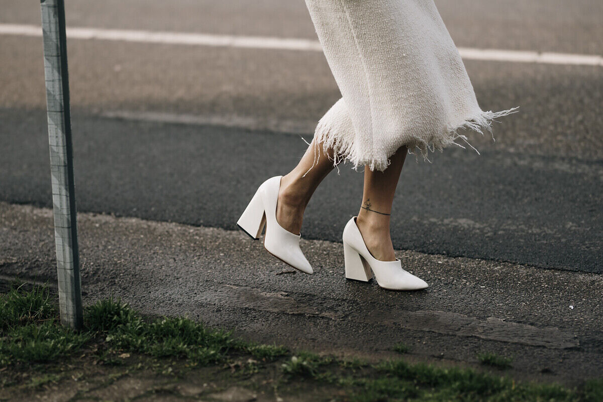 Clarks zwolnienia. Zdjęcie kobiety idącej ulicą, noszącej białą spódnicę midi z frędzlami na dole oraz białe czółenka na klockowym obcasie.