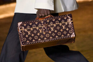 Zdjęcie z pokazu Louis Vuitton, prezentujące zbliżenie na brązowy kufer niesiony przez modela w białej marynarce i ciemnych spodniach. Prostokątny kufer jest pokryty słynnym monogramem luksusowego brandu i logo Louis Vuitton.