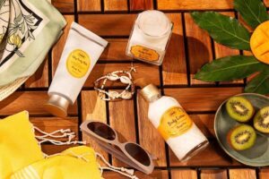 Zdjęcie kosmetyków Sabon, marki należącej do Yves Rocher. Na zdjęciu trzy produkty w białych opakowaniach z pomarańczowymi etykietami. Razem z nimi na drewnianym podłożu leżą okulary przeciwsłoneczne i bransoletka z muszelkami.