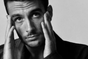 Czarno-białe zdjęcie Lorenzo Musetti, nowego ambasadora Bottega Veneta. Na fotografii sportowiec ma dwie ręce przy głowie, marszczy czoło i nosi elegancką marynarkę, pod którą ma koszulę.