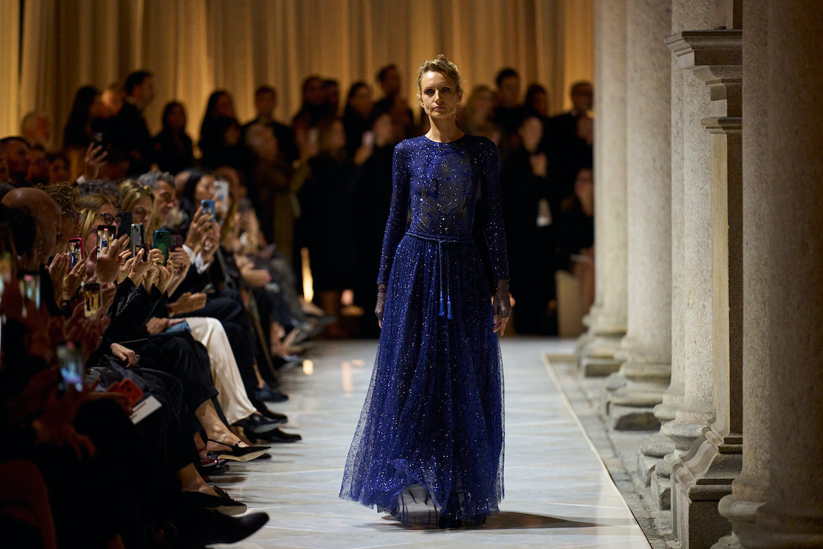 Modelka na wybiegu podczas pokazu Giorgio Armani w Mediolanie, ubrana w długą, połyskującą granatową suknię z długim rękawem, z publicznością fotografującą pokaz w tle.