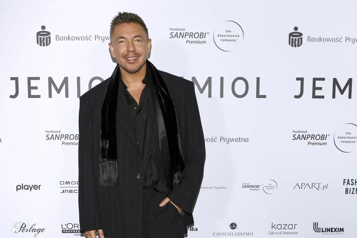 Łukasz Jemioł - polski projektant mody - na ściance po jubileuszowym pokazie. Jemioł ubrany jest w czarny garnitur. Na ściance widać loga patronów medialnych pokazu.