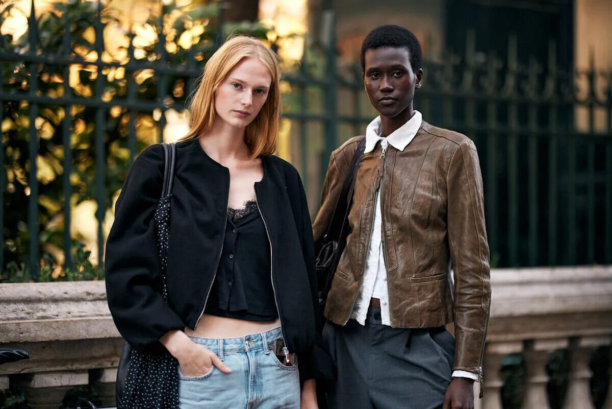 Najlepsze podcasty o modzie 2026. Na zdjęciu dwie osoby stoją na ulicy w miejskim otoczeniu, ubrane w minimalistyczne, casualowe stylizacje inspirowane modą codzienną. Jedna ma na sobie czarną kurtkę i jasne dżinsy, druga brązową skórzaną kurtkę i ciemne spodnie. Stylizacja nawiązuje do współczesnych trendów streetwearowych i naturalnej elegancji.