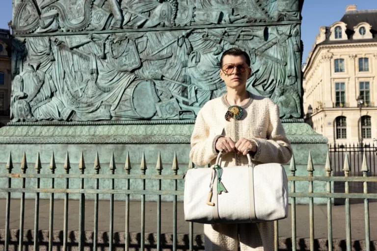 Adam Leja. Mężczyzna w jasnym kostiumie i okularach przeciwsłonecznych stoi przed monumentalną rzeźbą, trzymając dużą jasną torbę w dłoniach.