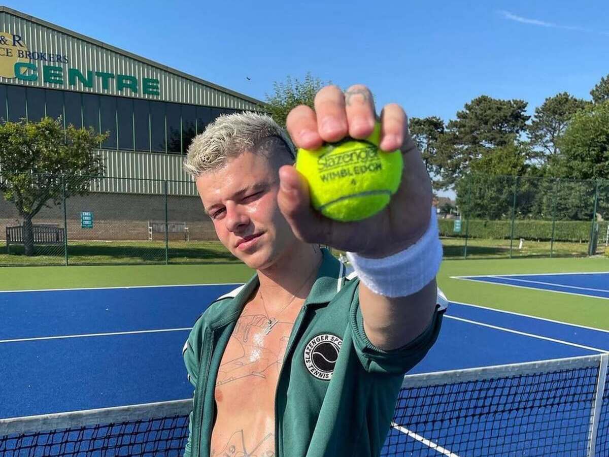 Alexei Hamblin Slazenger pokolenie Z. Młody mężczyzna na korcie tenisowym trzyma przed kamerą piłkę tenisową z napisem „Wimbledon”, w tle widać budynek i niebieskie niebo.