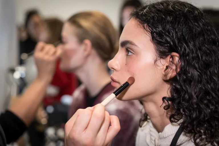 Gwen Stefani zamyka Gxve Beauty. Makijażystka nakłada róż pędzlem na policzek modelki podczas przygotowań backstage przed pokazem mody.