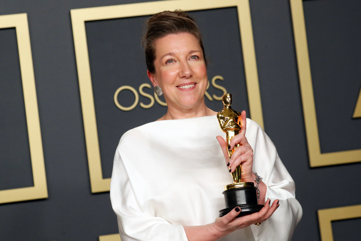 Jacqueline Durran kto to. Na zdjęciu: kostiumografka Wichrowych Wzgórz i Barbie. Jacqueline Durran pozuje z Oscarem w dłoniach na tle ścianki z napisem „Oscars”, ubrana w jasną, minimalistyczną suknię, uśmiecha się do aparatu.