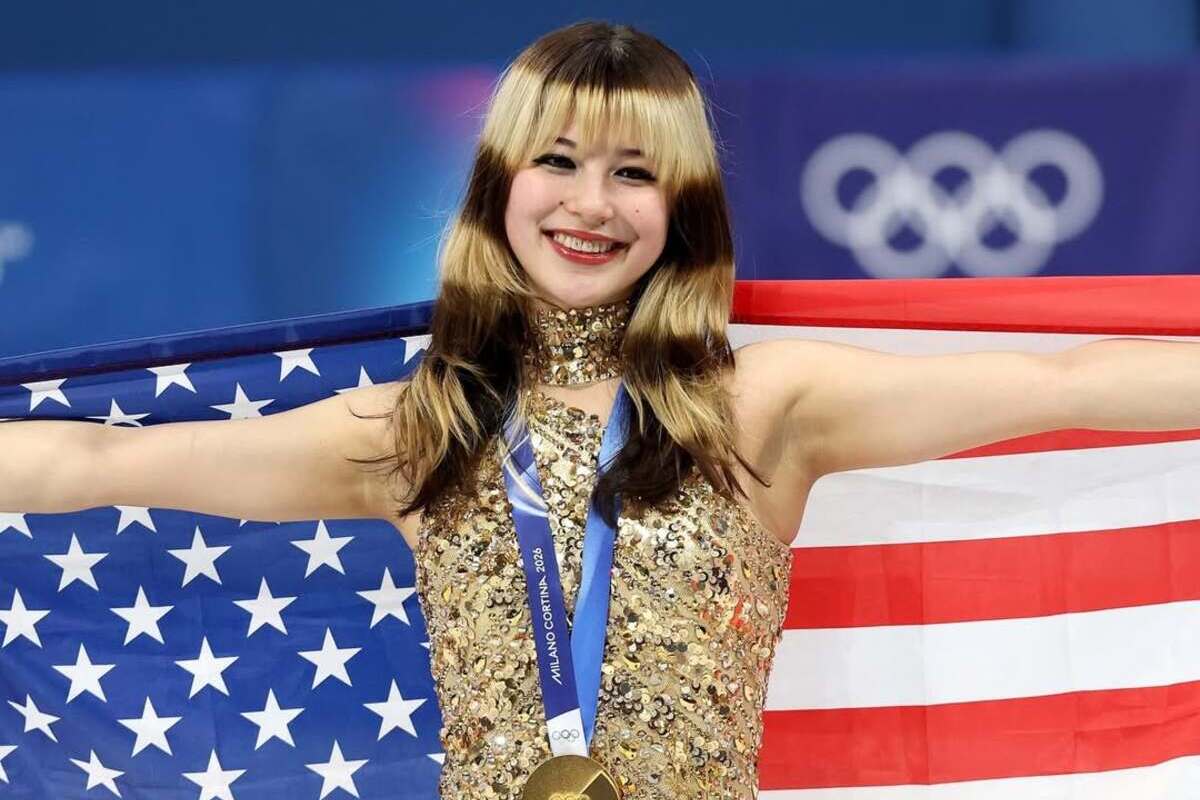 Styl Alysy Liu po olimpijskim występie widoczny jest w złotym kostiumie startowym, który łączy klasyczną elegancję z nowoczesną formą. Łyżwiarka stoi na lodzie z rozłożonymi ramionami, trzymając amerykańską flagę i prezentując złoty medal. Zdjęcie podkreśla emocje zwycięstwa oraz spójność jej sportowego i wizerunkowego przekazu.