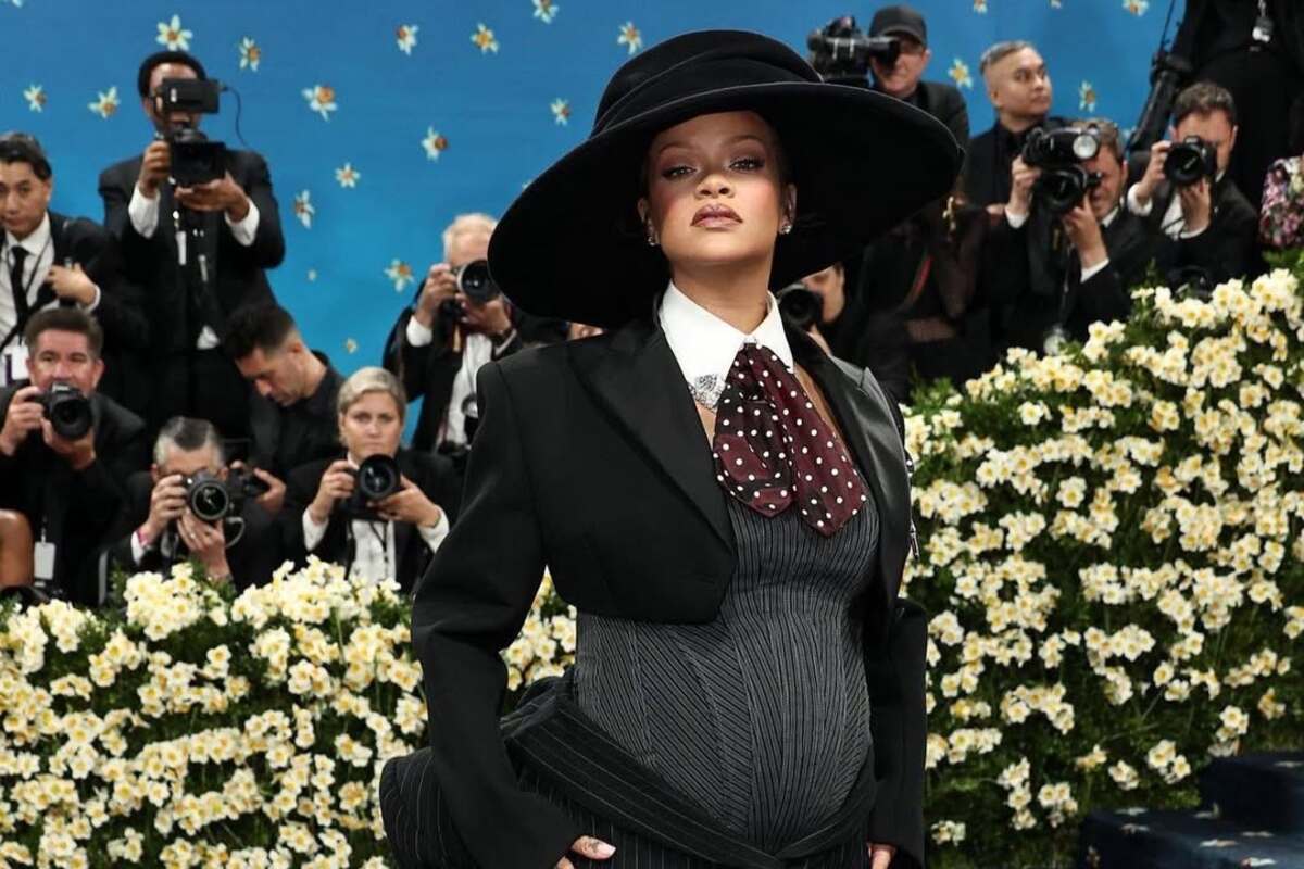 Met Gala dress code 2026. Na zdjęciu: Rihanna, uczestniczka Met Gali na czerwonym dywanie w czarnej stylizacji inspirowanej męskim tailoringiem, z szerokim kapeluszem i krawatem w grochy, pozująca na tle fotografów i kwiatowej scenografii.