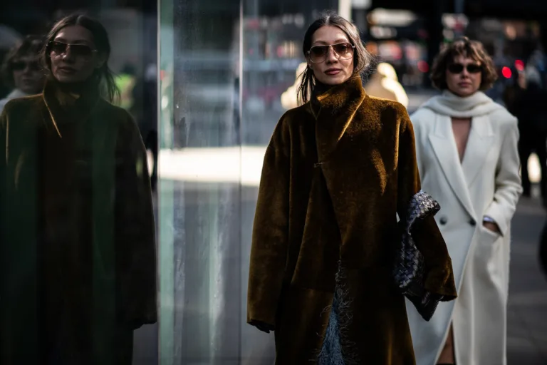 Certyfikat B Corp co to. Dwie elegancko ubrane kobiety idą ulicą w miejskiej scenerii, prawdopodobnie w centrum dużego miasta. Na pierwszym planie znajduje się kobieta w długim, brązowym płaszczu o wysokim kołnierzu, w okularach przeciwsłonecznych, trzymająca pod pachą czarną, plecioną kopertówkę; jej sylwetka odbija się w szklanej witrynie po lewej stronie kadru. Za nią idzie druga kobieta ubrana w jasny, kremowy płaszcz i szalik, również w ciemnych okularach. Tło jest lekko rozmyte, z widocznymi światłami i przechodniami, co podkreśla miejski, modowy charakter fotografii.