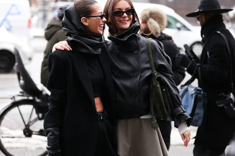 Ekspansja Sinsay. Dwie stylowo ubrane kobiety w czarnych płaszczach i okularach przeciwsłonecznych idą ulicą w zimowej scenerii, obejmując się i uśmiechając, a w tle widać przechodniów oraz zaparkowane rowery.