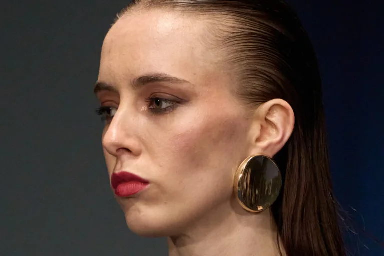Ozempic a rynek biżuterii. Zbliżenie na twarz kobiety z zaczesanymi do tyłu włosami, w dużym, okrągłym złotym kolczyku i czerwonej szmince, sfotografowane w półprofilu na ciemnym tle.