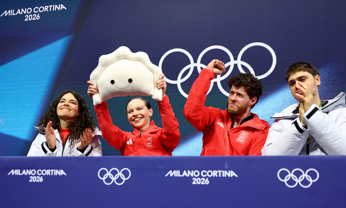 Pluszowy pieróg igrzyska olimpijskie. Cztery osoby w strojach reprezentacji siedzą na scenie z logo Milano Cortina 2026 i olimpijskimi kołami w tle, świętując podczas konferencji. Jedna z nich unosi nad głową maskotkę przypominającą pluszowy pieróg, co nadaje wydarzeniu lekki, radosny charakter. Atmosfera igrzyska podkreślona jest przez gesty triumfu i oklaski uczestników.
