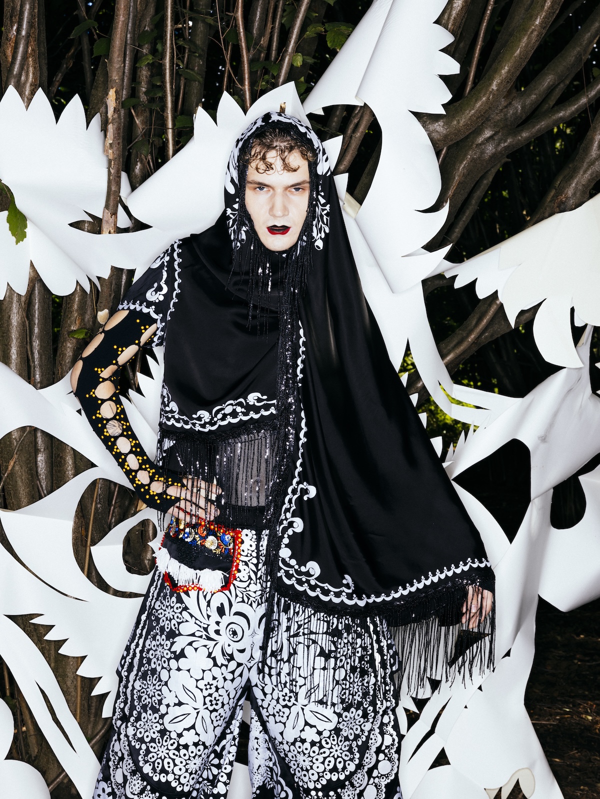 Model stoi wśród drzew na tle białych, wycinanych form przypominających ptaki i kwiaty. Ma na sobie czarny, haftowany strój z frędzlami i wzorami inspirowanymi folklorem oraz nakrycie głowy z czarną chustą.