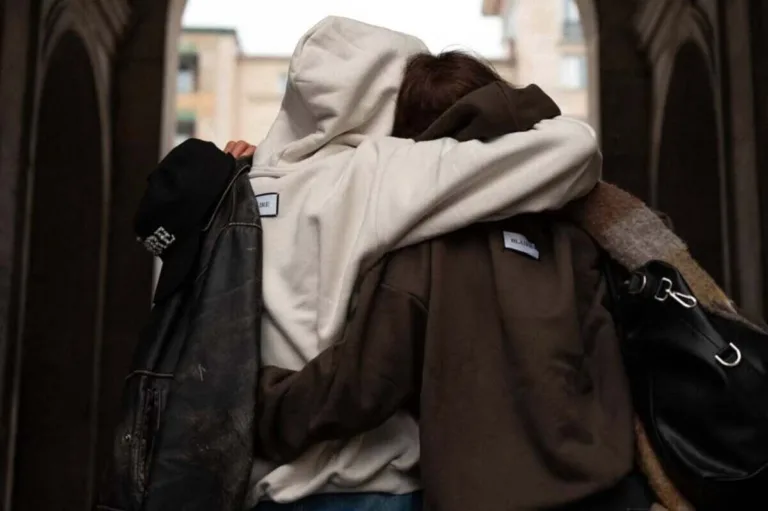 Dwie osoby stoją tyłem do aparatu, obejmując się, ubrane w oversize’owe bluzy BLÁIRE afera. BLÁIRE w kolorach beżowym i brązowym, na tle miejskiej architektury.