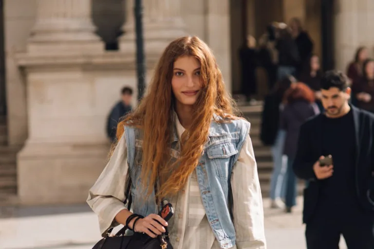 Zdjęcie przedstawia stylową kobietę o długich, miedzianych włosach, która stoi na placu, uśmiechając się do aparatu. Ma na sobie beżową koszulę w paski, jeansową kamizelkę, szerokie jeansy typu baggie i czarno-białe buty typu sneakers na platformie. W jednej ręce trzyma czarną torbę z widocznym brelokiem. Ten nowoczesny i modny look idealnie wpisuje się w trendy modowe. Jeśli posiadasz w szafie podobne perełki, dowiedz się, gdzie sprzedać ubrania online.