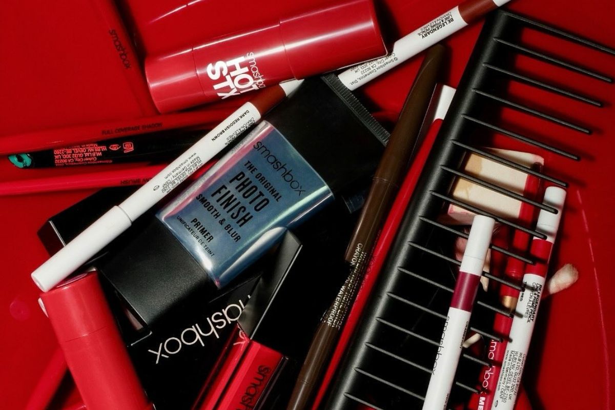 Zdjęcie przedstawia artystyczny kolaż profesjonalnych produktów do makijażu marki Smashbox, ułożonych na intensywnie czerwonym tle, gdzie w centrum uwagi znajduje się kultowa baza Photo Finish Smooth & Blur, otoczona przez czerwone szminki, błyszczyki, kredki do ust i pędzle, co przywołuje na myśl marki kosmetyczne, które zniknęły z Polski, a ich brak wciąż odczuwają fanki wysokiej jakości wykończenia i trwałości.