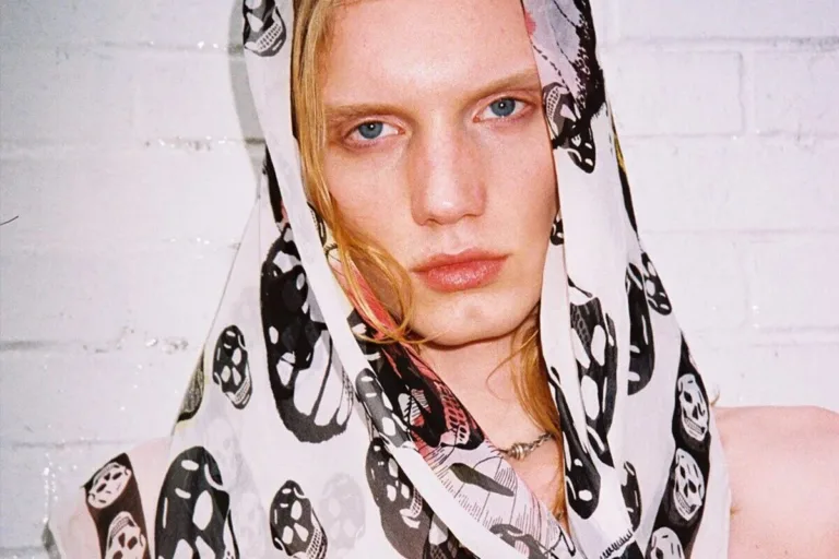 Szalik w czaszki Alexander McQueen. Modelka w zbliżeniu portretowym ma na głowie lekko zarzuconą, półtransparentną jedwabną chustę Skull Scarf w kolorze ecru z czarnym motywem czaszek, która miękko opada wokół twarzy, kontrastując z jasną skórą i surowym, minimalistycznym tłem.