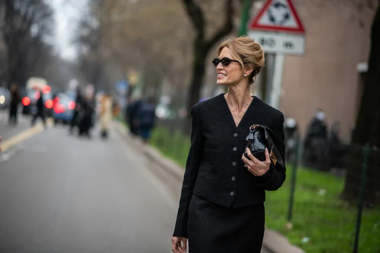 Petycja o dodatkowy dzień wolny za święto w niedzielę. Elegancko ubrana kobieta w czarnym kostiumie spaceruje ulicą, trzymając w dłoni małą czarną torebkę; ma upięte włosy i okulary przeciwsłoneczne, a w tle widać rozmyte sylwetki ludzi oraz znak drogowy.