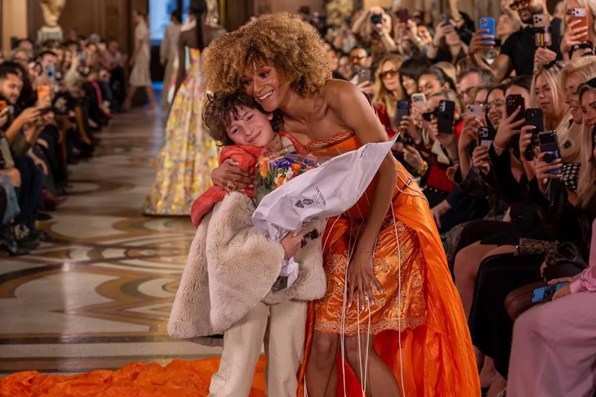 10-letni Max Alexander - najmłodszy projektant w historii Paris Fashion Week. Na zdjęciu chłopiec z bukietem kwiatów obejmowany na wybiegu przez modelkę w pomarańczowej sukni.