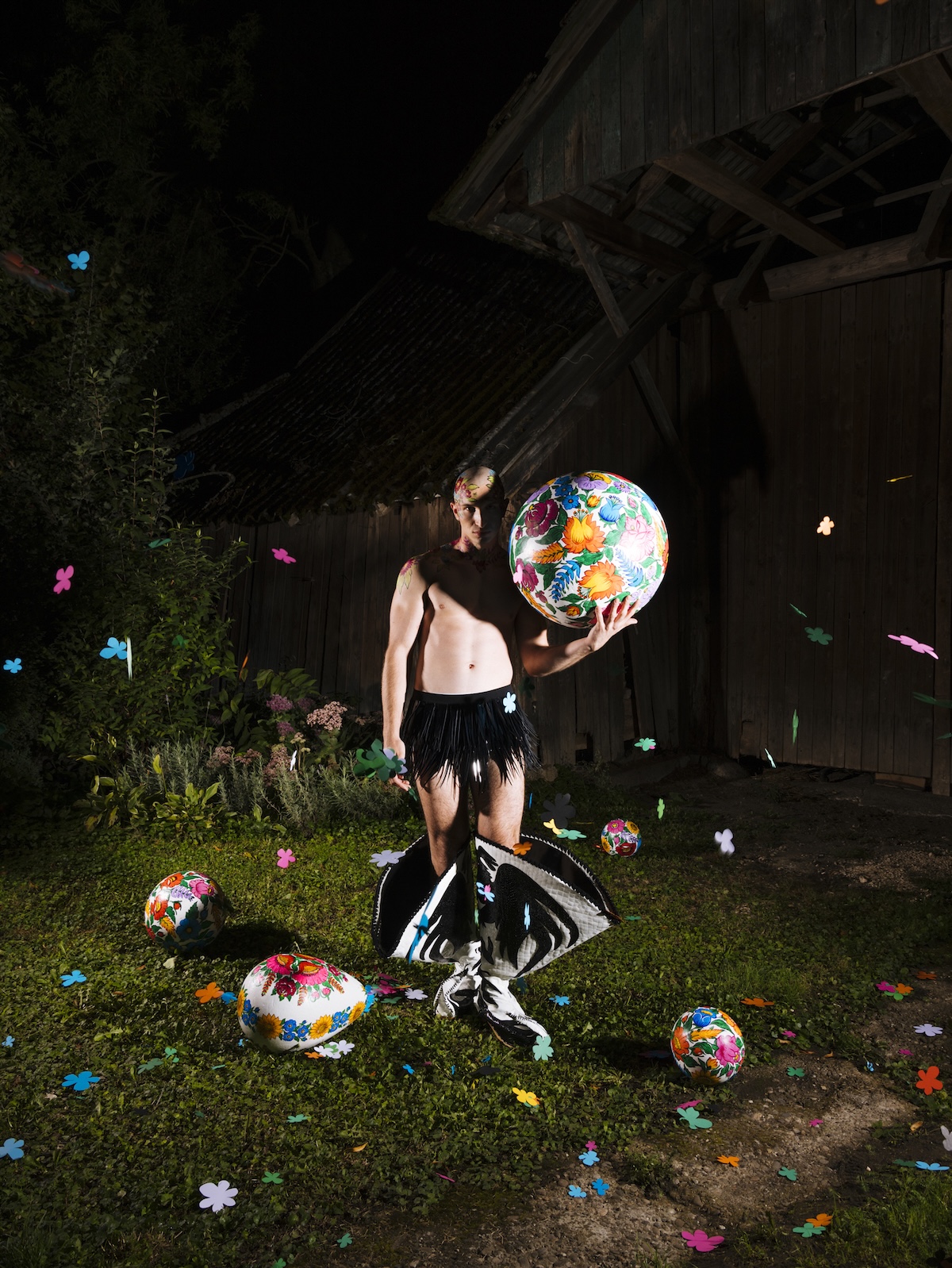 Model stoi nocą przed drewnianym budynkiem, trzymając dużą kulę z kwiatowym wzorem. Wokół leżą dekoracyjne, malowane jajka i kolorowe papierowe kwiaty, a model ma na sobie czarną spódnicę z frędzlami i wysokie buty.
