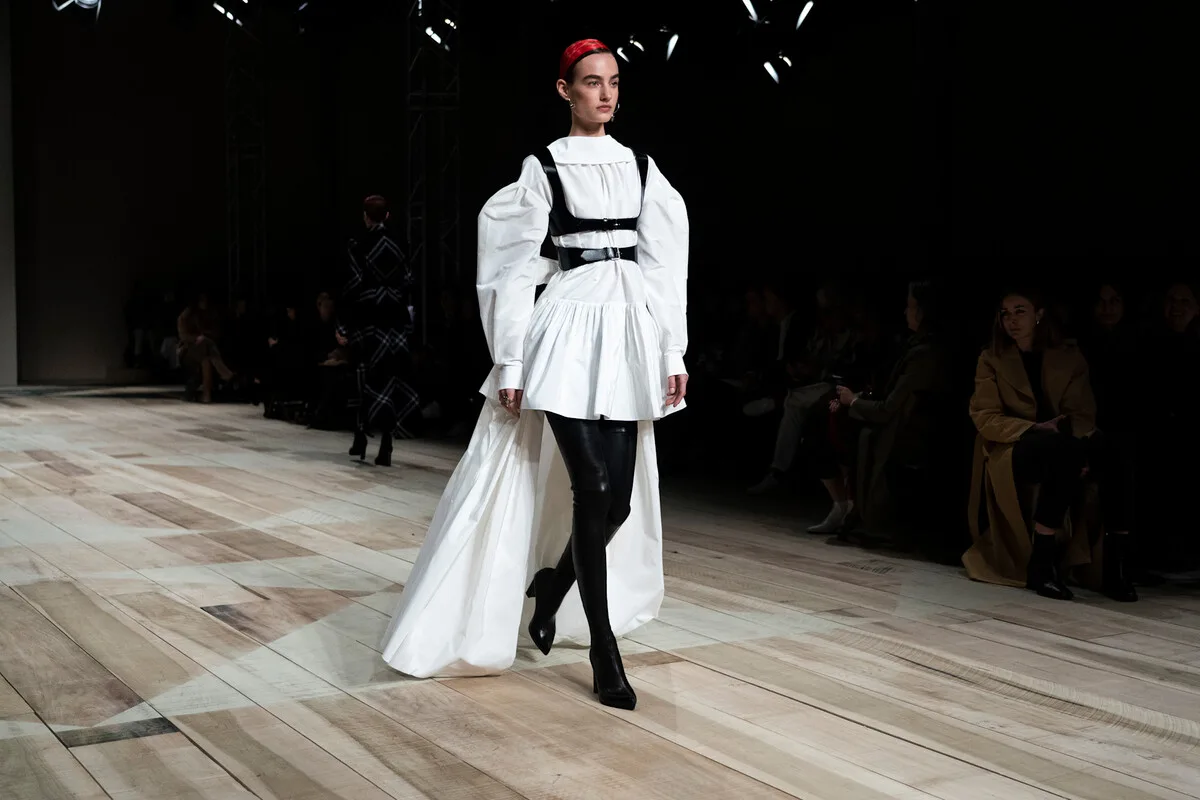 Zwolnienia w Alexander McQueen 2026. Modelka idzie po wybiegu podczas pokazu mody, prezentując białą suknię o rozbudowanej formie z bufiastymi rękawami i długim trenem. Stylizację uzupełniają czarne pasy przypominające uprząż oraz czarne, dopasowane buty i rajstopy. Scena odbywa się na jasnym drewnianym wybiegu w ciemnej sali, z widownią siedzącą po bokach.