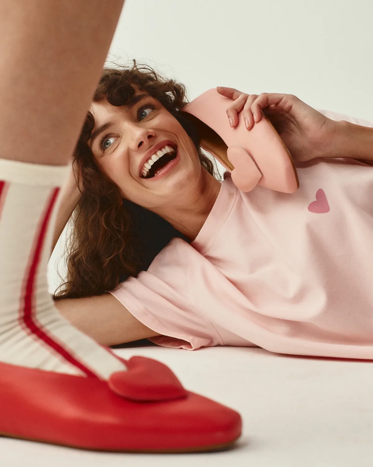 Uśmiechnięta modelka w różowej stylizacji prezentuje baleriny z kolekcji Balagan x She is Sunday. Kadr ma lekki, zabawny charakter i podkreśla dziewczęcą estetykę projektu. Pastelowe kolory kontrastują z intensywną czerwienią obuwia.