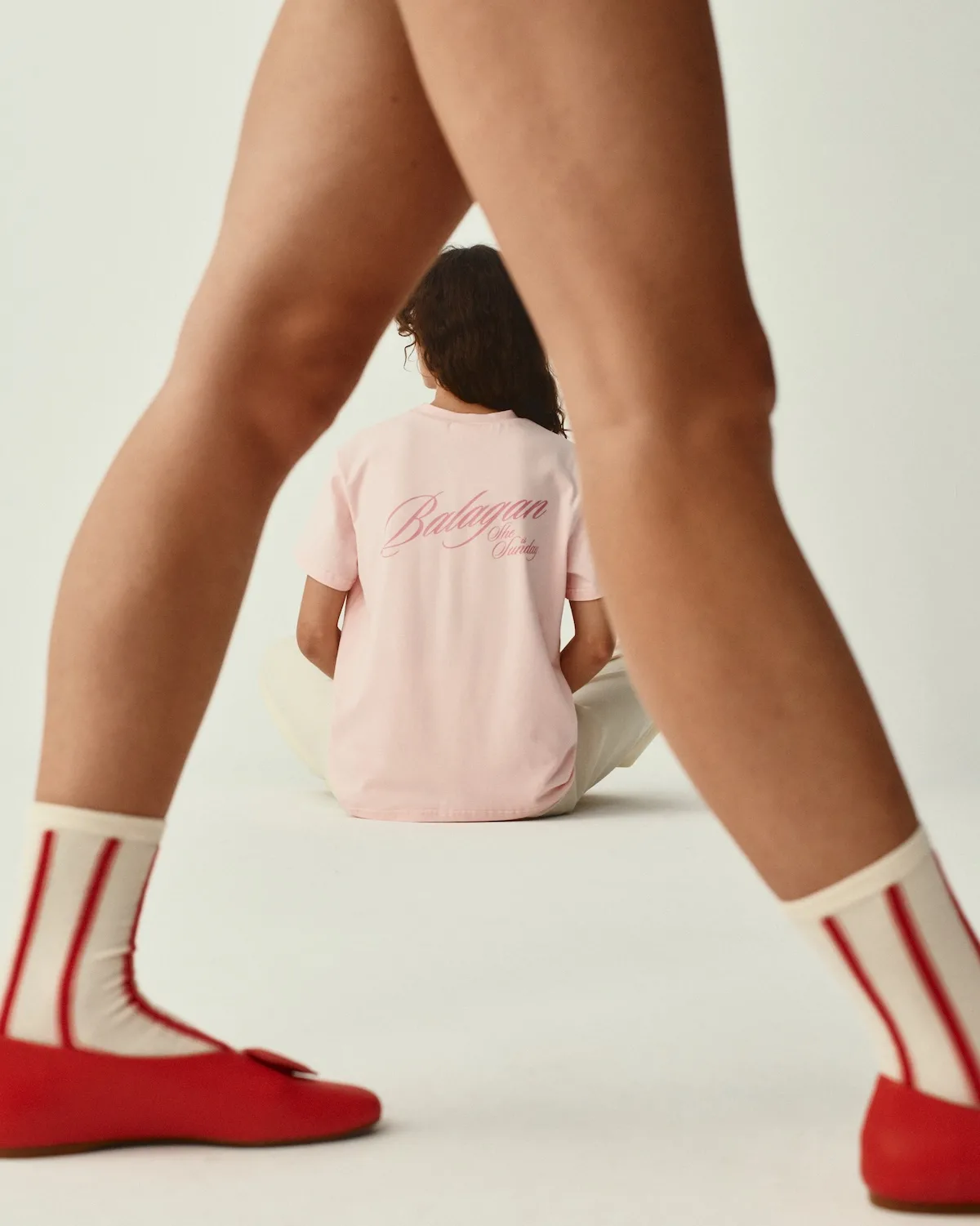 Modelka w czerwonych balerinach i skarpetkach w paski stoi na pierwszym planie, tworząc dynamiczną kompozycję. W tle widoczna jest druga osoba w koszulce z logo Balagan x She is Sunday. Zdjęcie podkreśla charakterystyczny styl kolekcji i jej graficzną spójność.