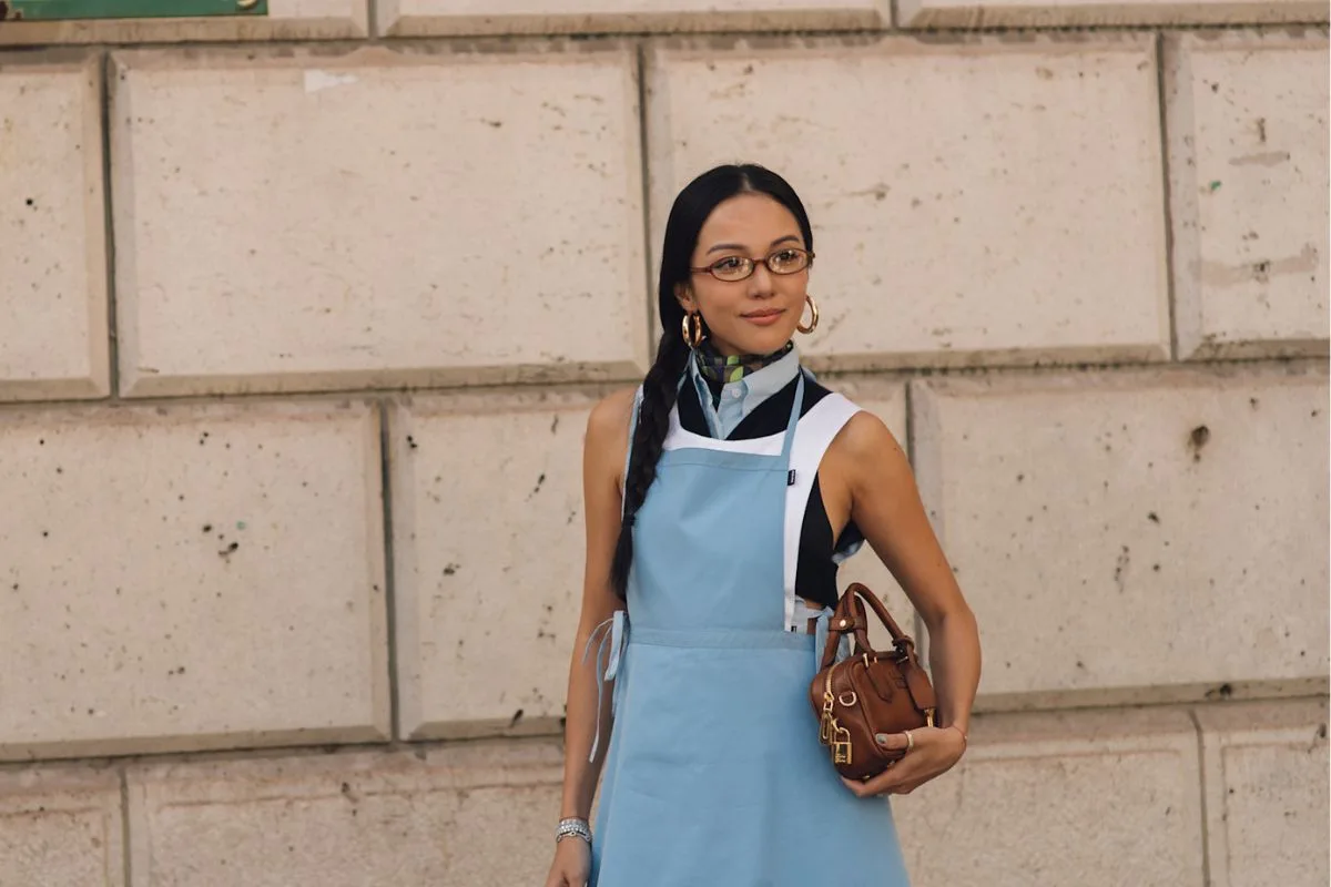 Kobieta w błękitnej sukience typu fartuszek i okularach na tle kamiennej ściany w Paryżu – inspiracja na wiosenne stylizacje oraz informacja, czy w majówkę 2026 są otwarte sklepy.