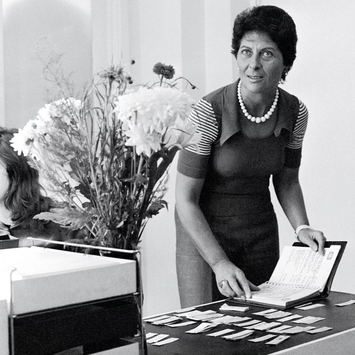 Czarno-białe zdjęcie Joan Burstein przy pracy w butiku Browns w Londynie. Projektantka i kuratorka mody przegląda notatki oraz selekcjonuje kolekcje – scena oddaje jej intuicyjne podejście do odkrywania nowych talentów.