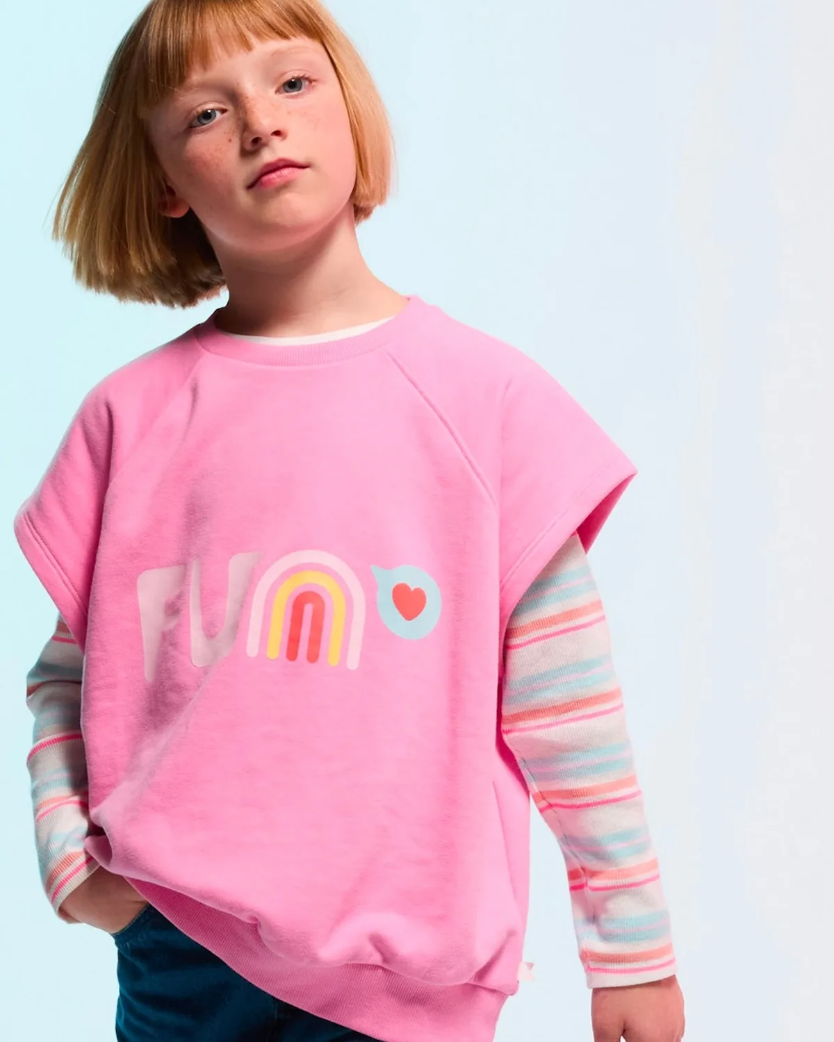 Dziewczynka w różowej bluzie z kolorowym nadrukiem i długim rękawem w paski prezentuje casualowy strój dziecięcy. Stylizacja jest lekka, kolorowa i dopasowana do codziennego użytku. Fotografia oddaje charakter oferty modowej dla dzieci, którą oferowała marka Okaïdi.