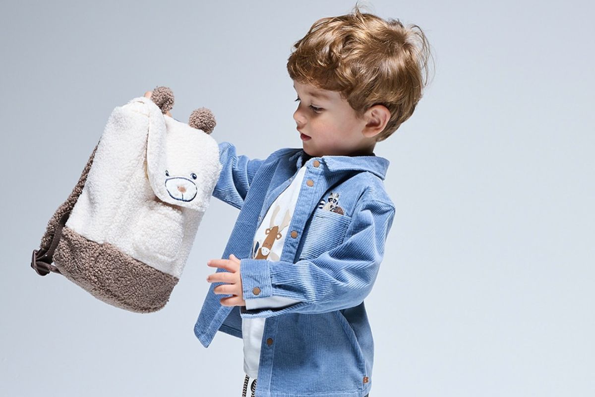 Okaidi upadłość Polska. Chłopiec w niebieskiej koszuli i białym T-shircie ogląda dziecięcy plecak w kształcie misia. Stylizacja ma casualowy charakter i nawiązuje do oferty odzieży oraz akcesoriów dla najmłodszych. Zdjęcie ilustruje asortyment typowy dla marek z segmentu mody dziecięcej.
