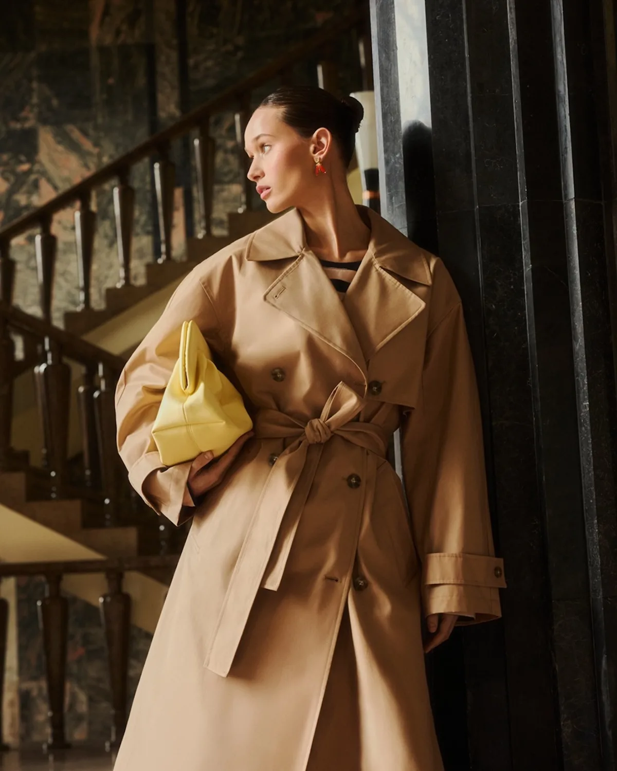 Modelka prezentuje klasyczny beżowy trencz, który podkreśla kierunek w stronę ponadczasowej, bardziej premium oferty – spójny z strategicznym rozwojem segmentu wyższej jakości. Stylizacja z kontrastową, żółtą torebką odzwierciedla jednocześnie potrzebę przyciągania uwagi klientów, co w kontekście wyników finansowych wiąże się z rosnącymi wydatkami marketingowymi i walką o sprzedaż.