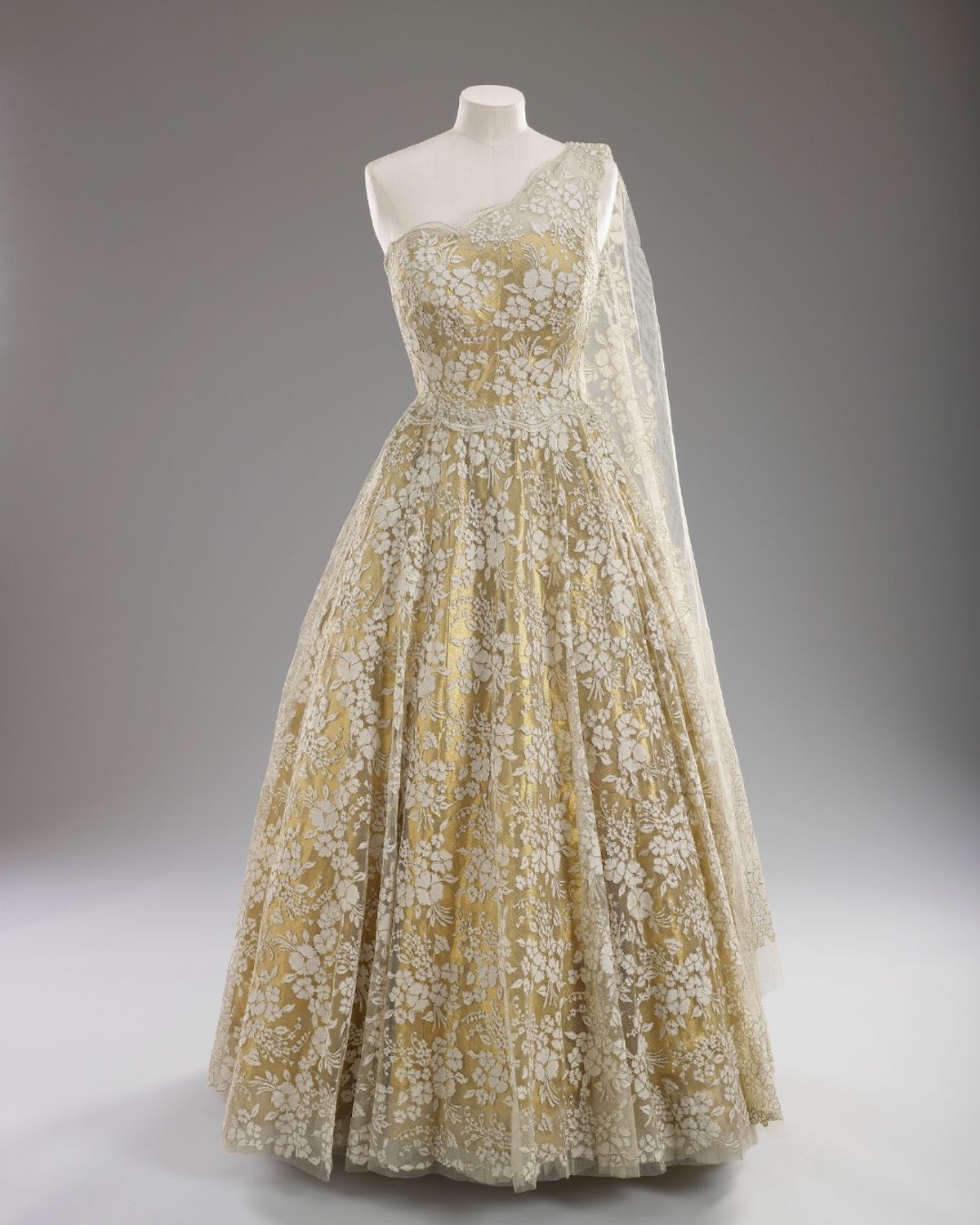 Jedna z wieczorowych sukni królowej Elżbiety II – bogato zdobiona, z haftem i delikatną koronką, podkreślająca ceremonialny charakter królewskich wystąpień. Na wystawie w Pałacu Buckingham tego typu projekty pokazują, jak istotną rolę odgrywały tkaniny i rzemiosło w budowaniu prestiżu wizerunku Elżbiety II.