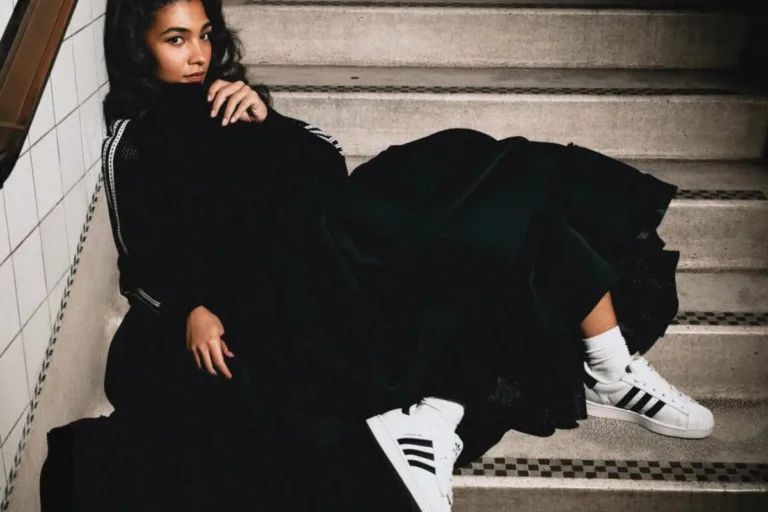 Kobieta siedzi na schodach w miejskiej przestrzeni, ubrana w czarną, luźną stylizację o eleganckim charakterze. Na nogach ma białe sneakersy adidas Superstar z charakterystycznymi trzema paskami, które kontrastują z ciemnym outfitem. Stylizacja łączy modę uliczną z klasyczną estetyką, podkreślając ponadczasowy design butów.