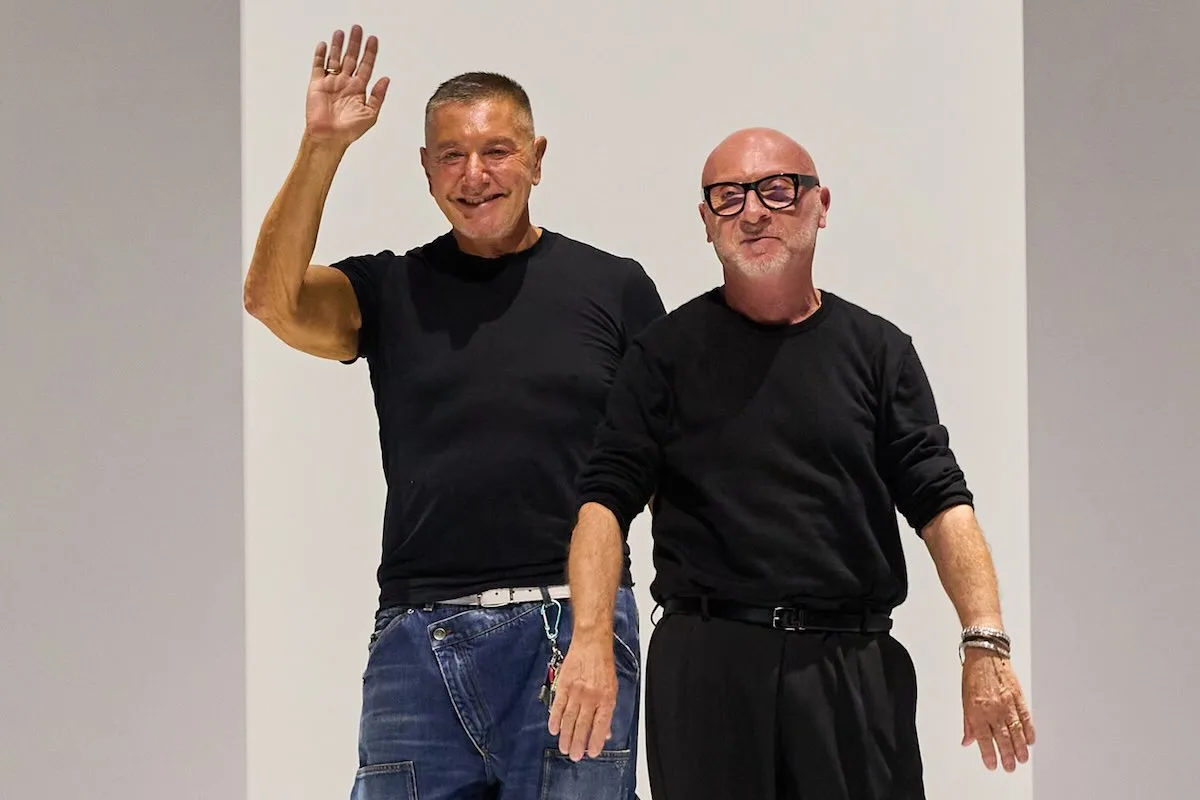 Stefano Gabbana i Domenico Dolce - twórcy Dolce & Gabbana, stoją obok siebie na wybiegu, ubrani w czarne koszulki; jeden z nich macha do publiczności po pokazie mody.