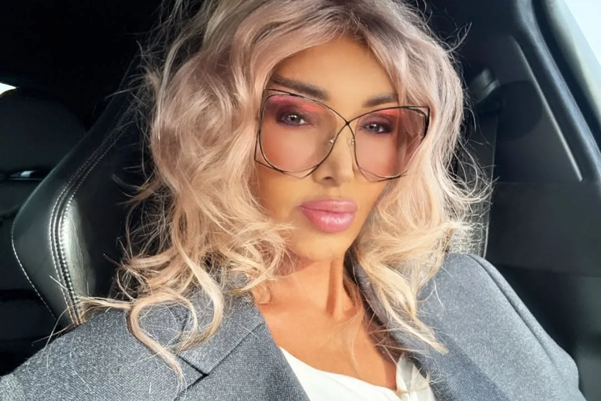 Ewa Minge Epstein. Kobieta w okularach przeciwsłonecznych i szarym płaszczu siedzi w samochodzie, wykonując selfie.