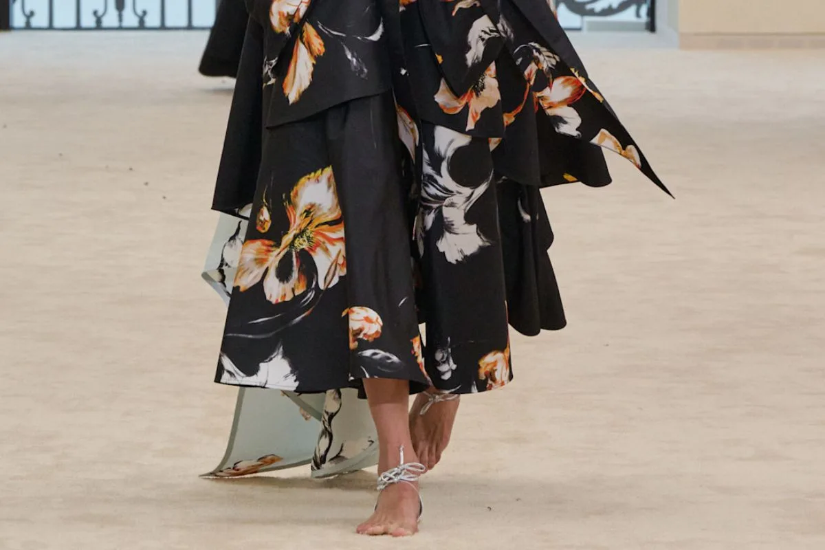 Modelka na wybiegu w zwiewnej, kwiatowej spódnicy, prezentująca minimalistyczne sandały z samą piętą Chanel, które niemal znikają na stopie. Konstrukcja ograniczona do cienkiego wiązania i elementu przy pięcie tworzy efekt bosej stopy. To właśnie te sandały z samą piętą Chanel wywołały dyskusję o „butach bez butów”.