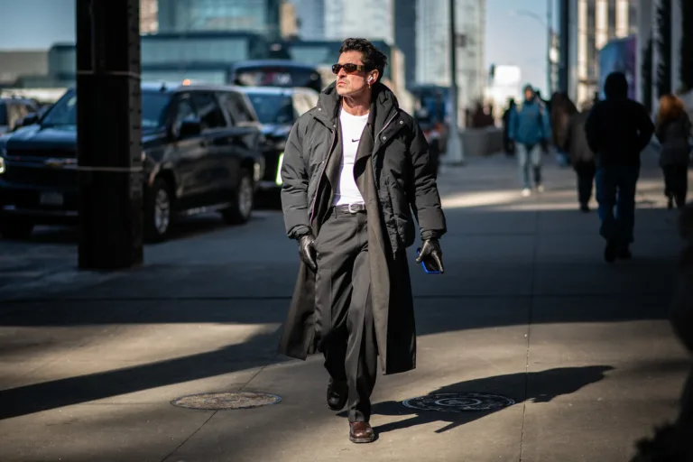 Zarzuty UOKiK promocje Black Red White, Peek & Cloppenburg, Żabka Nano. Mężczyzna w ciemnym płaszczu i okularach przeciwsłonecznych idzie chodnikiem w centrum miasta, trzymając w ręku telefon. W tle widać uliczny ruch i przechodniów, co podkreśla miejską, dynamiczną scenerię. Zdjęcie ma charakter ilustracyjny i nawiązuje do tematyki handlu oraz codziennych zakupów.