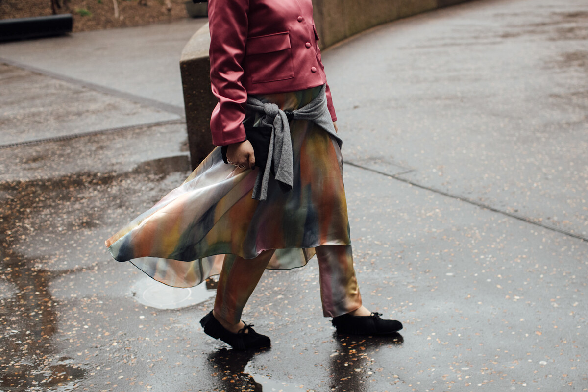 Zwolnienia unilever bydgoszcz. Osoba w kolorowej, półprzezroczystej spódnicy i różowej kurtce przechodzi przez mokrą ulicę, niosąc czarną torebkę.