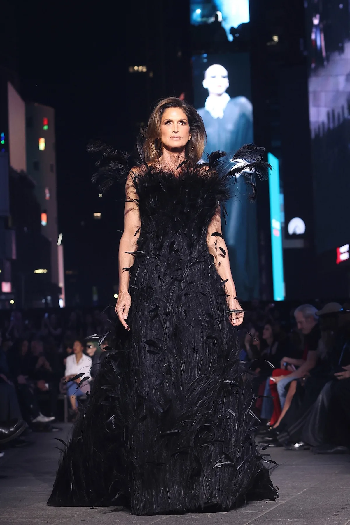 Cindy Crawford na wybiegu podczas pokazu Gucci Resort PO S27 w długiej czarnej sukni wieczorowej zdobionej piórami i transparentnymi detalami.