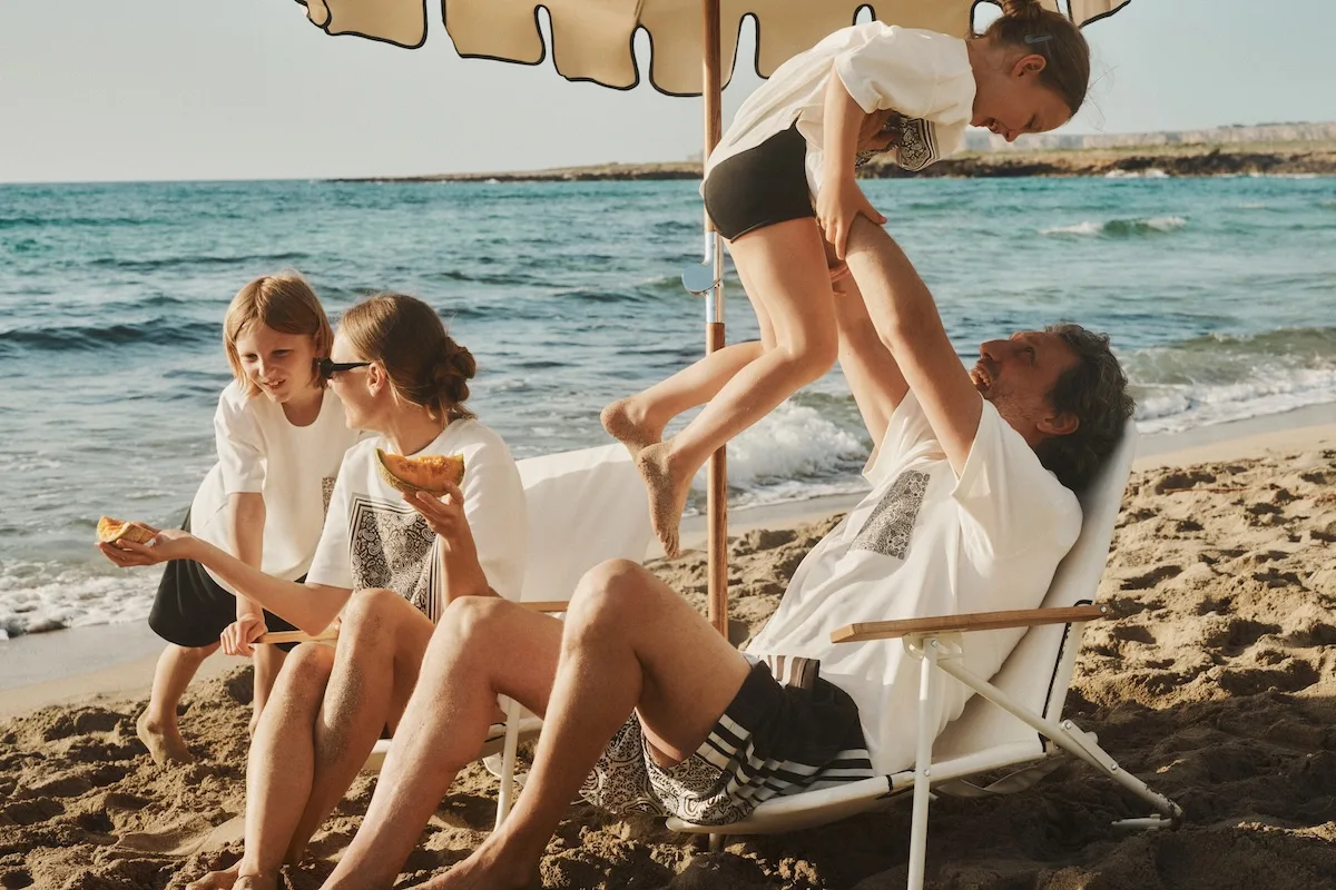 Modele z dziećmi odpoczywają na plaży w ubraniach z kolekcji LEBRAND x RESERVED. Zdjęcie utrzymane jest w swobodnym, wakacyjnym klimacie.