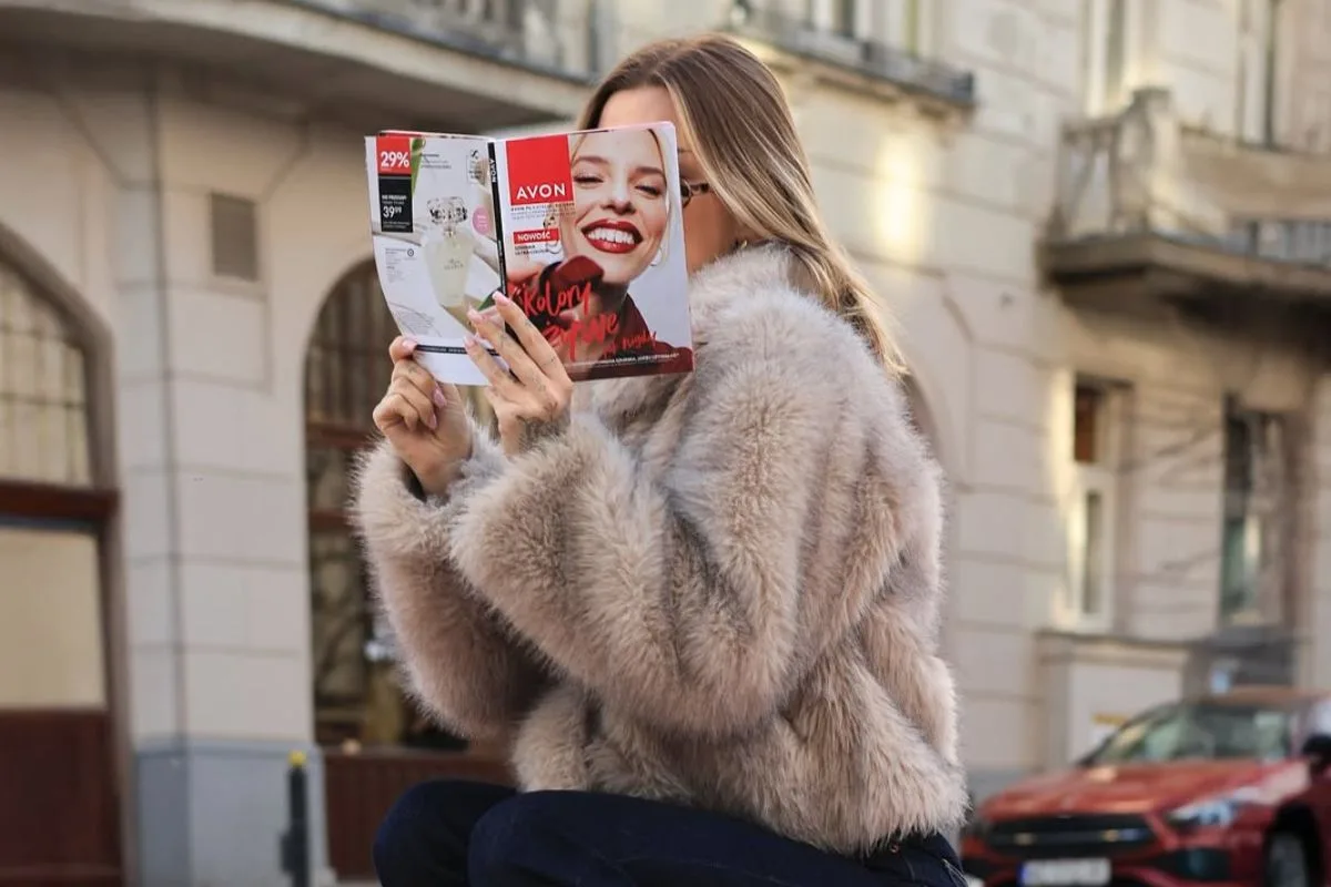 Avon UOKiK kara. Kobieta siedzi na ulicy i trzyma przed twarzą katalog Avon. W tle widać miejską zabudowę, a zdjęcie nawiązuje do sprzedaży kosmetyków i działalności marki Avon.