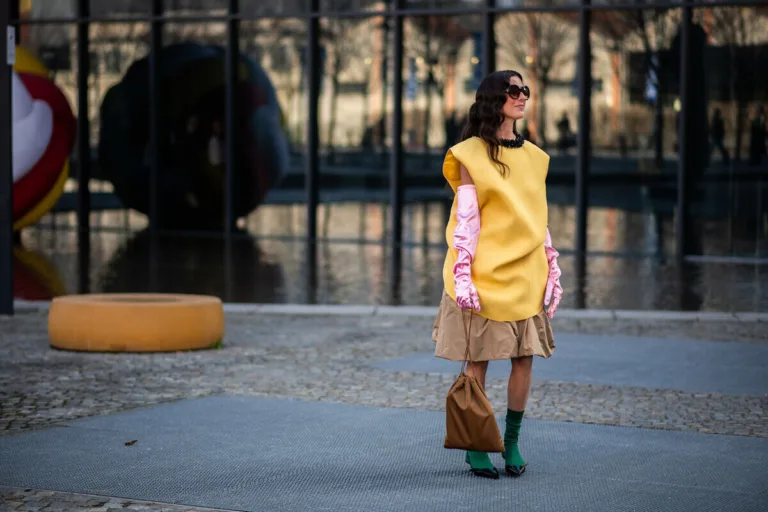 Kobieta prezentuje odważną stylizację street style podczas tygodnia mody, łącząc oversize'ową żółtą tunikę z różowymi rękawiczkami i zielonymi dodatkami. Zdjęcie nawiązuje do świata fashion, w którym platformy e-commerce, takie jak ABOUT YOU Marketplace, otwierają markom modowym nowe możliwości dotarcia do klientów w Polsce i Europie.