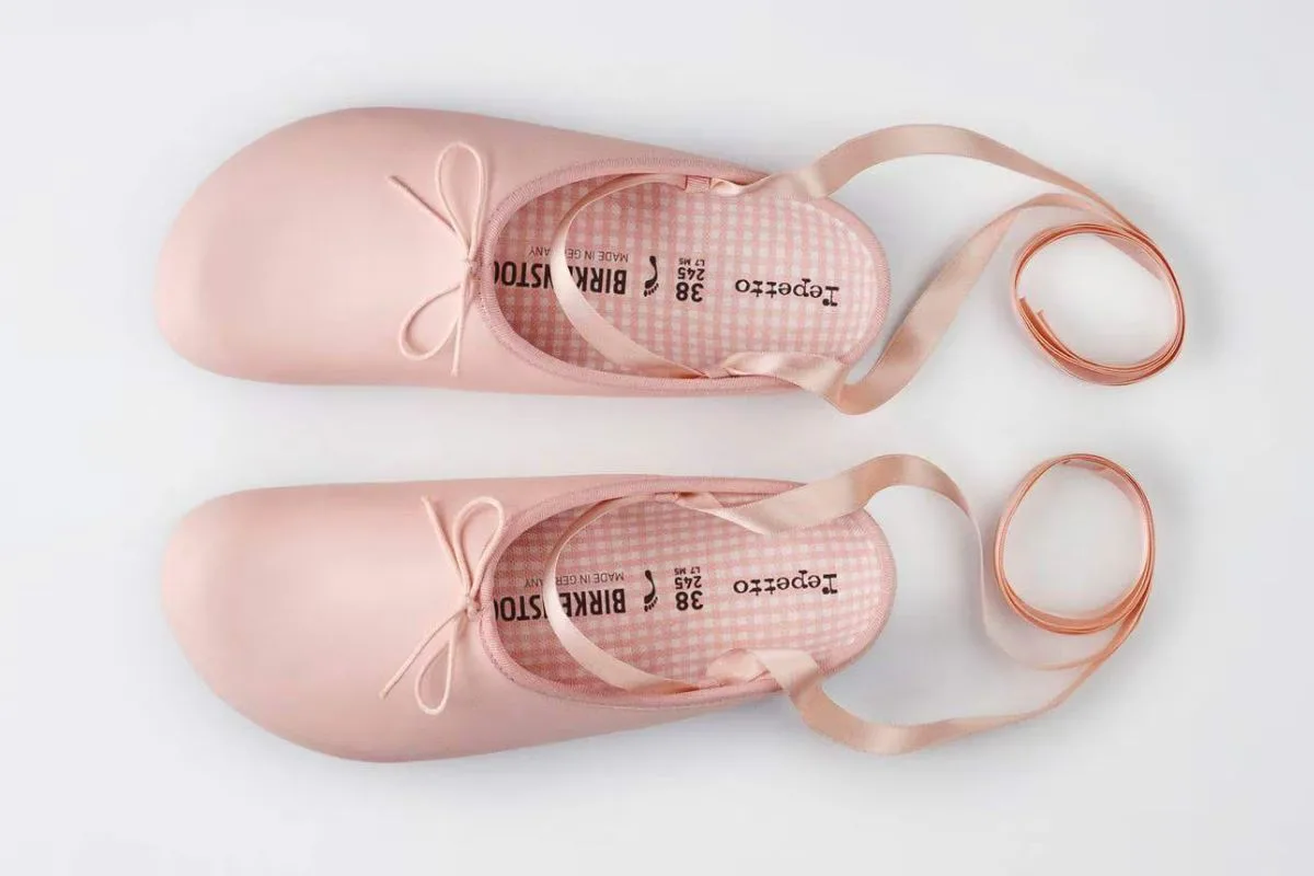 Birkenstock x Repetto. Różowe buty z zaokrąglonymi noskami, kokardkami i długimi satynowymi wstążkami, ułożone na jasnym tle.
