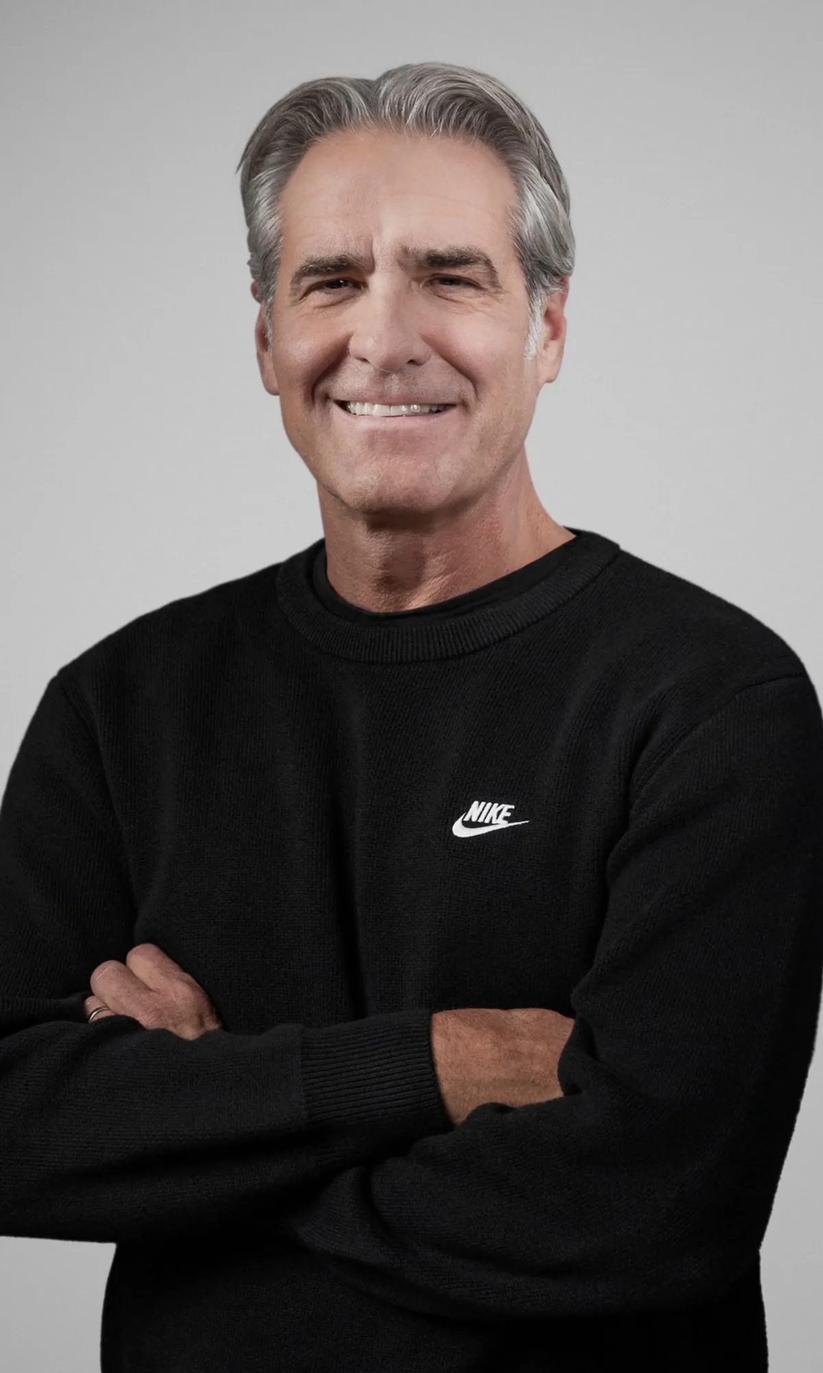 Elliott Hill, prezes Nike, pozuje w czarnym swetrze z logo marki na piersi. Menedżer stoi ze skrzyżowanymi rękami na tle jasnej, neutralnej ściany. W 2025 roku znalazł się na czele rankingu najlepiej opłacanych prezesów firm modowych i handlowych.